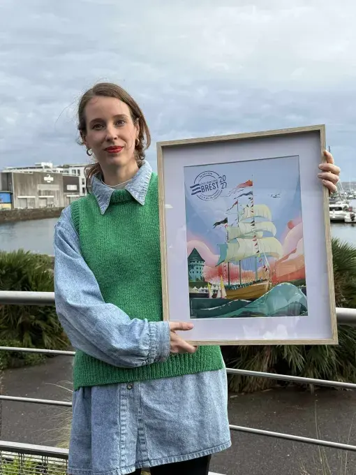 Une femme debout tient un cadre avec l'affiche des Fêtes maritimes de Brest 2027.