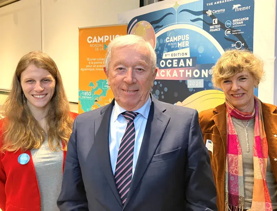 Un homme entouré de deux femmes, devant l'affiche présentant le campus mondial de la mer