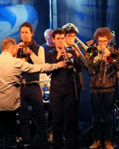 Photo montrant un groupe de 10 musiciens jazz jouant sur une scène en concert (saxophone, trombone, trompette) 