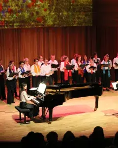 Photo montrant une vingtaine de choriste chantant sur scène accompagnée par une pianiste et dirigés par une cheffe de choeur.