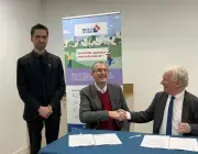 Deux hommes assis derrière une table se serrent la main. Un homme se tient debout à côté.