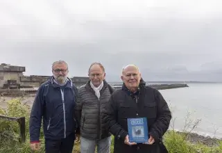 Trois hommes posent devant un bâtiment militaire. 