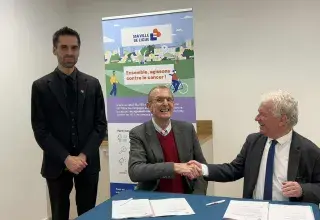 Deux hommes assis derrière une table se serrent la main. Un homme se tient debout à côté.